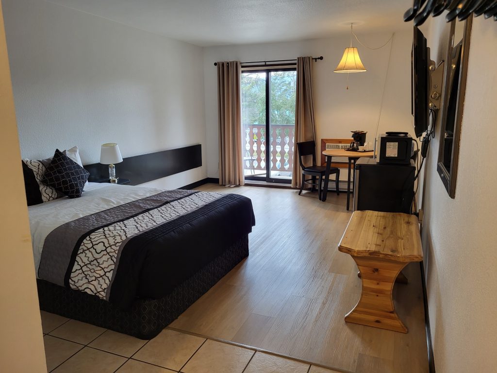 Single queen bed room with clean wood flooring, a side table and fridge, with glass sliding door.